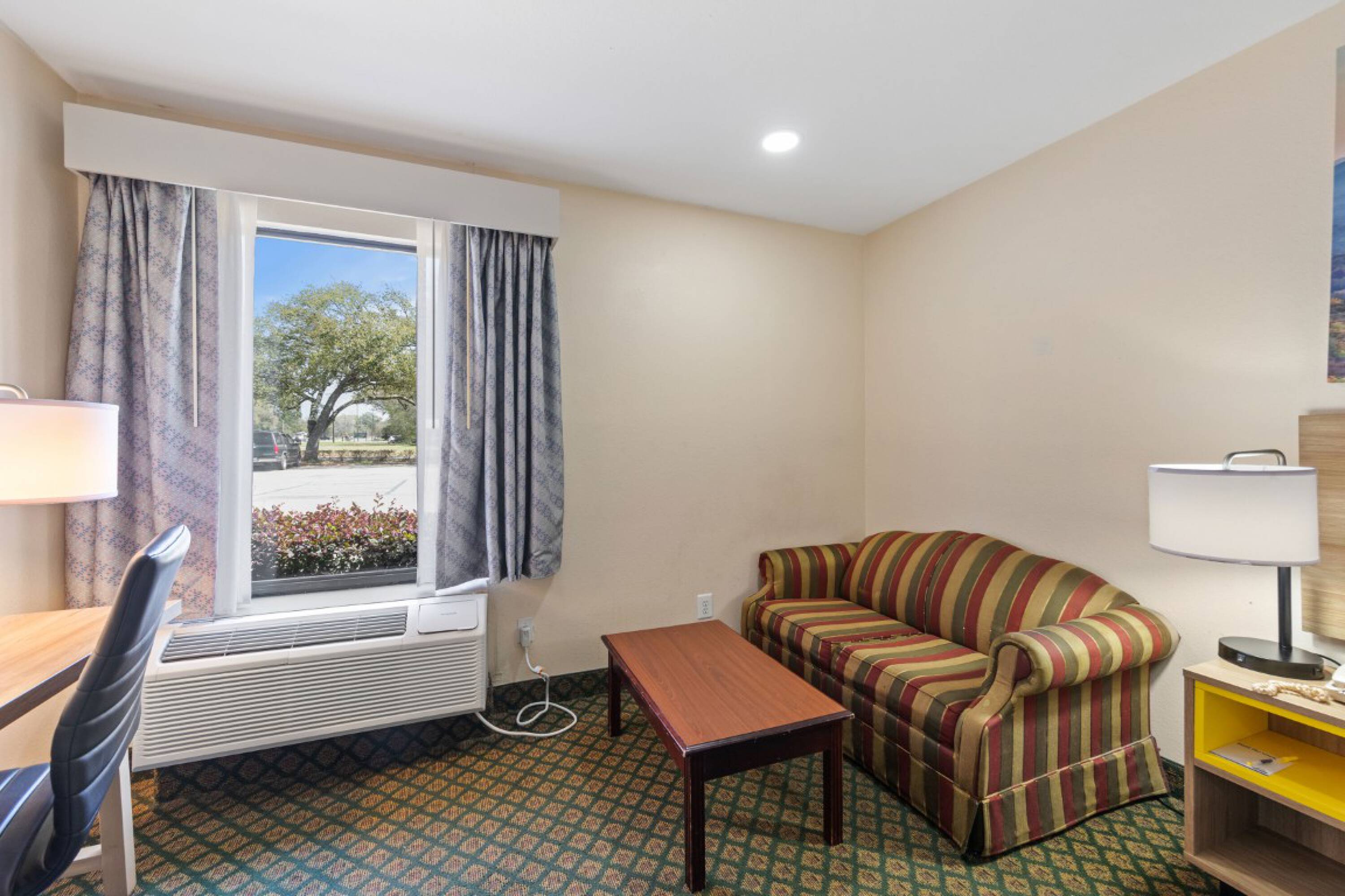 Cozy hotel room with a large window, striped sofa, wooden coffee table, work desk, and two table lamps at Days Inn & Suites by Wyndham Eunice.