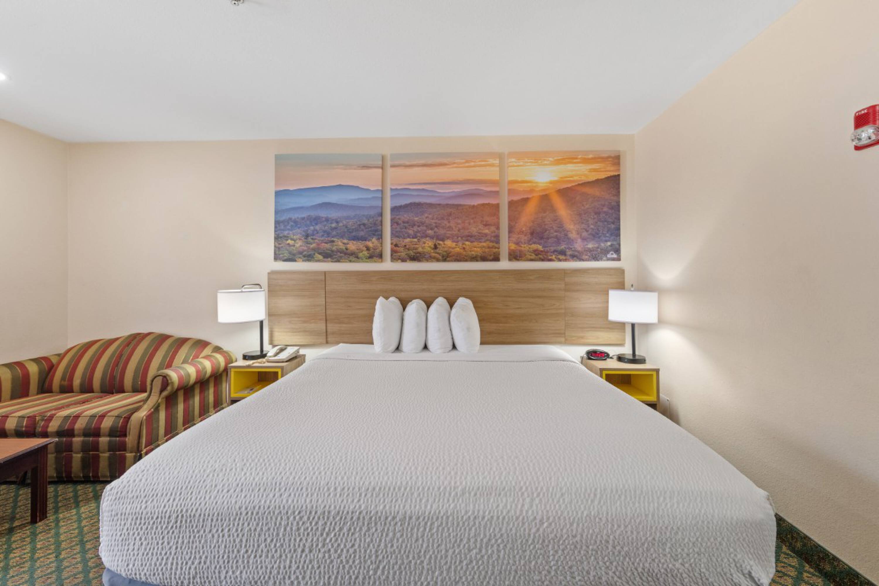 A well-appointed hotel room at Days Inn & Suites by Wyndham Eunice featuring a queen bed, two nightstands with lamps, a striped sofa, and scenic wall art.