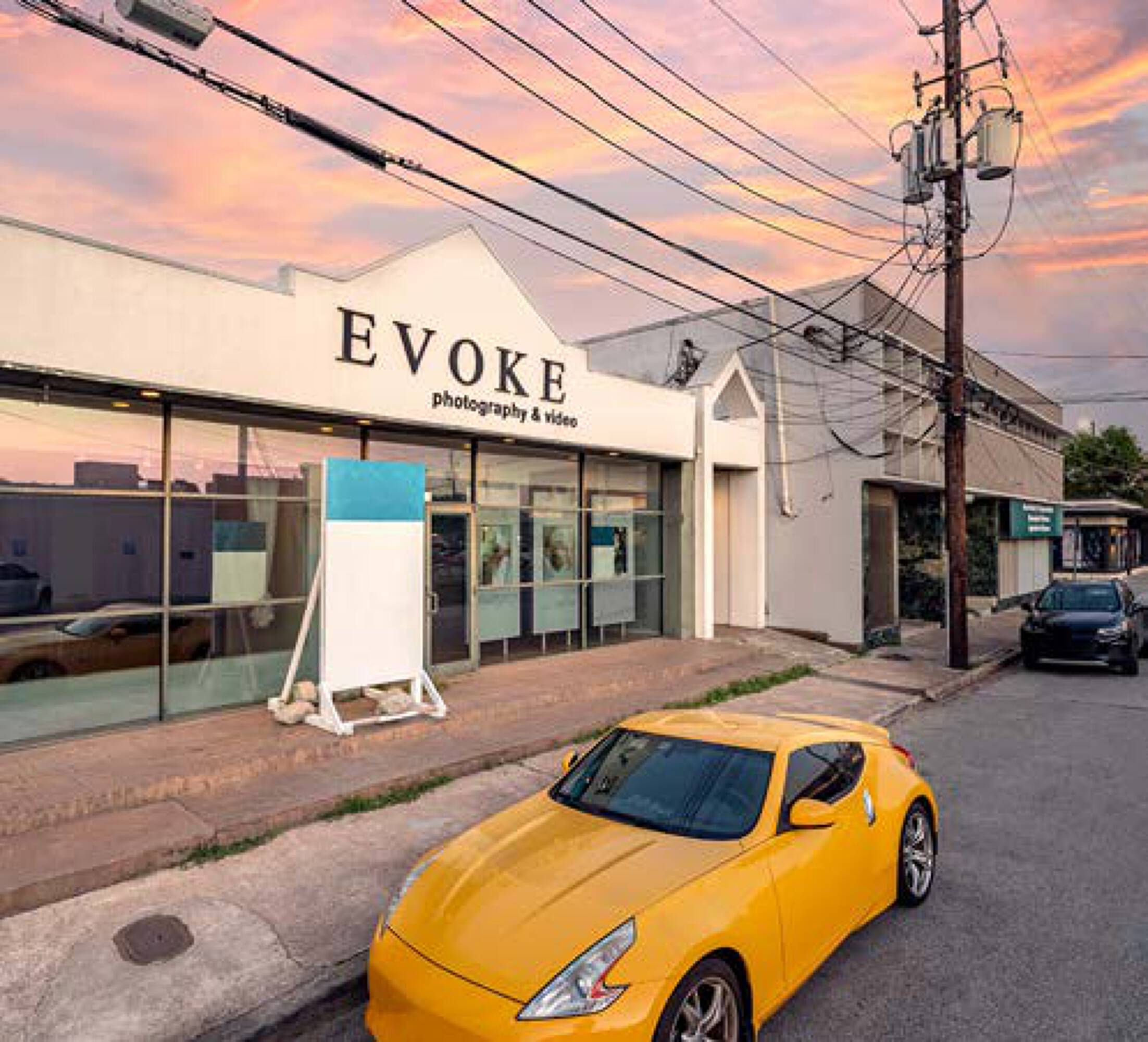 Bright yellow car parked in front of a modern retail/office building with large windows and "Evoke Photography & Video" signage.