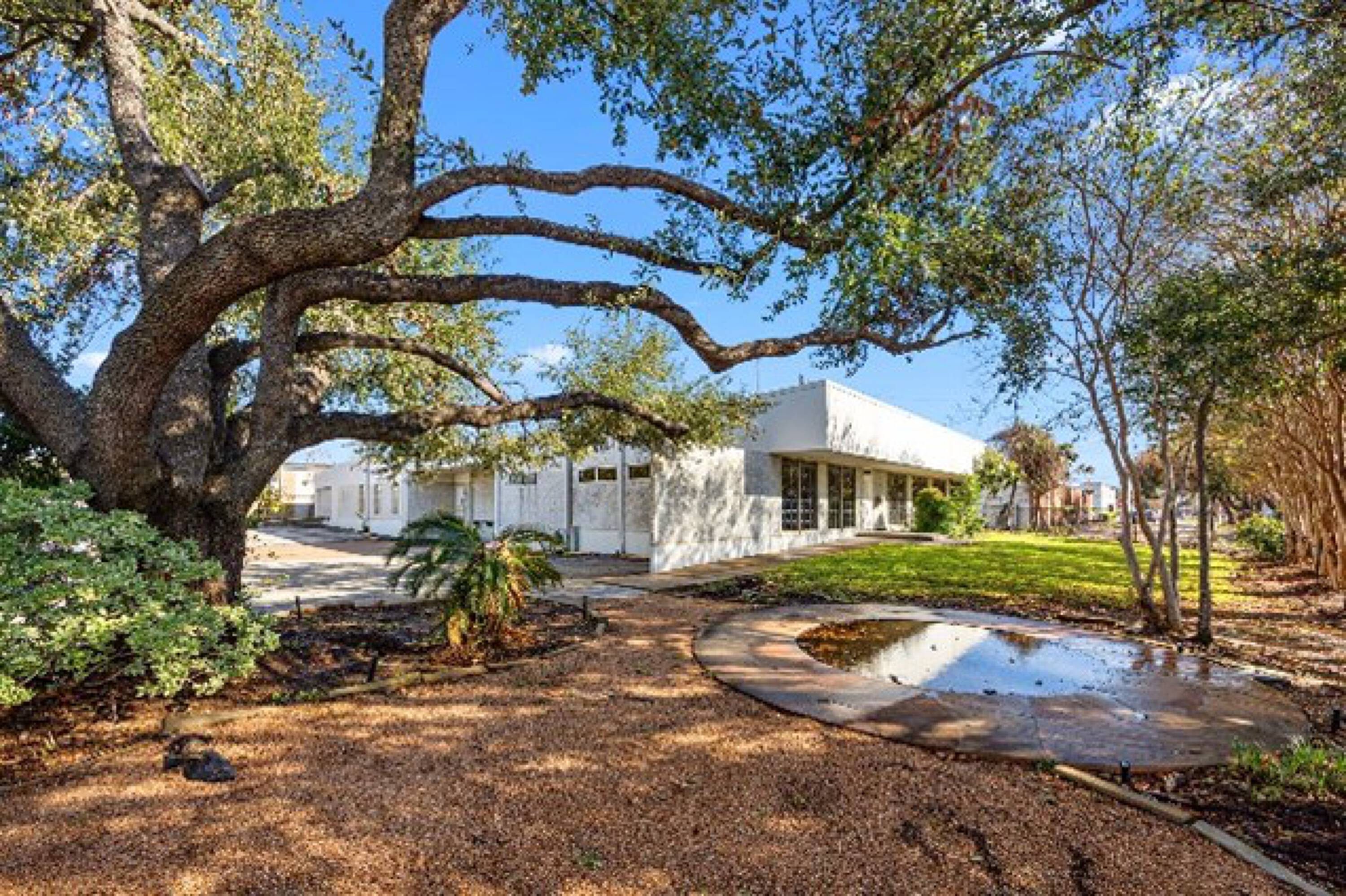 White industrial building with large windows and a landscaped front yard featuring a large tree and a fountain.