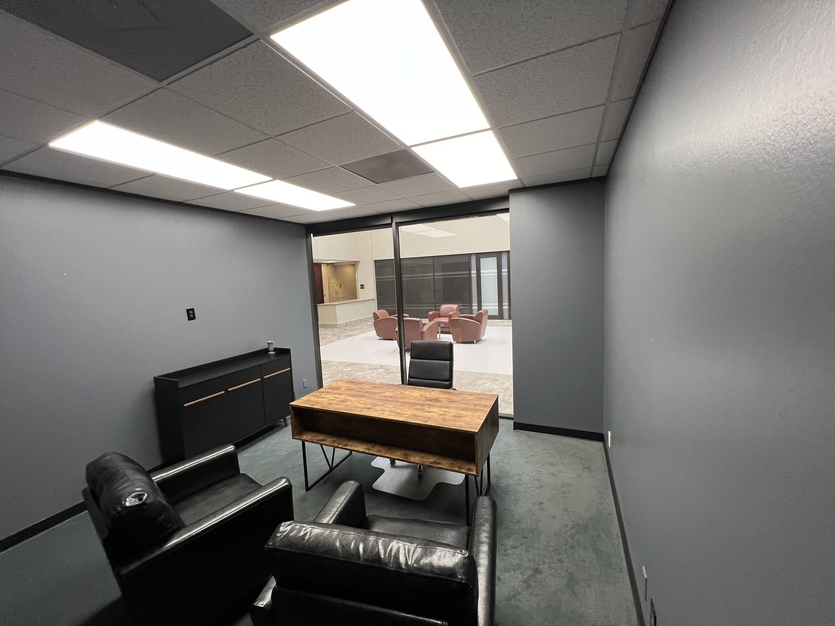 Office space with a wooden desk, black leather chairs, and large glass door providing natural light.