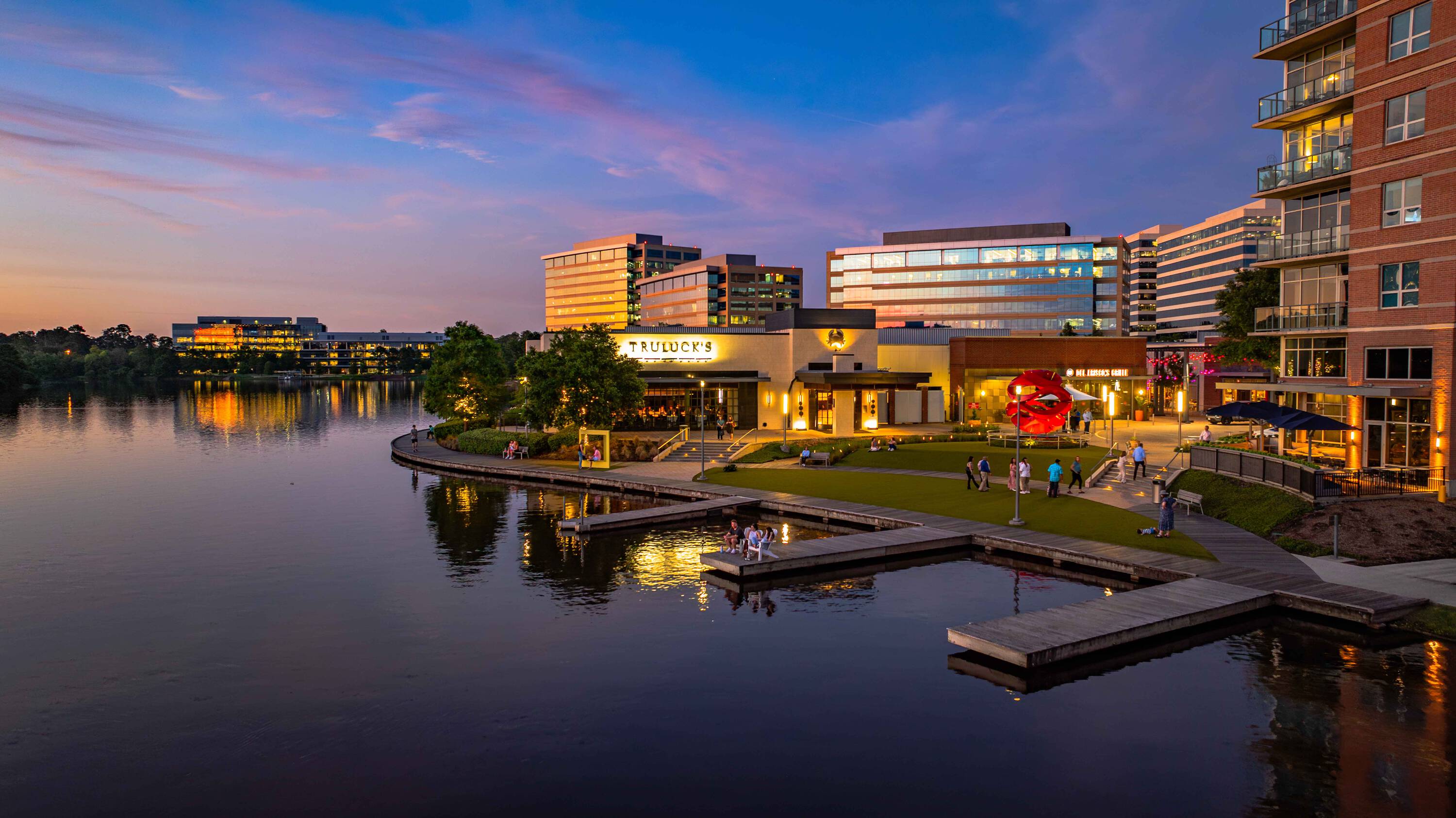 Evening view of Hughes Landing on Lake Woodlands with modern retail buildings, waterfront access, and pedestrian walkways.